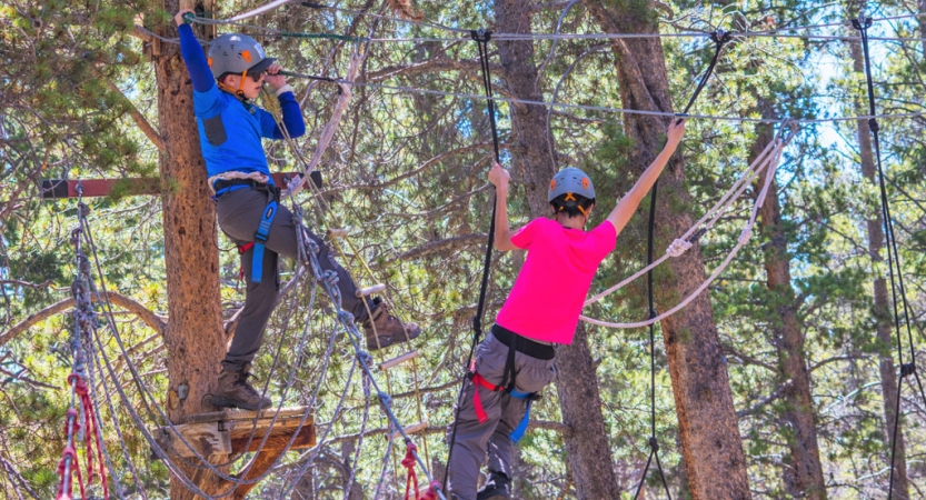 Two students wearing safety gear are secured by ropes as they navigate a high ropes course. 